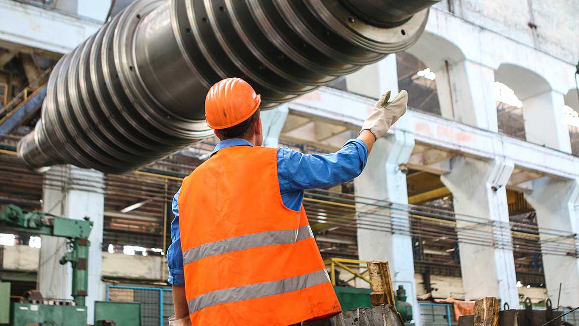 A man in an orange vest and hard hat standing under a large metal object.