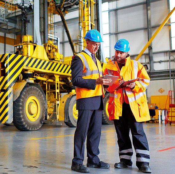 Two men in hard hats and yellow jackets standing next to a construction truck.