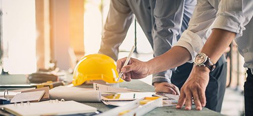 A man writing on paper while another person wearing hard hat looks at papers.